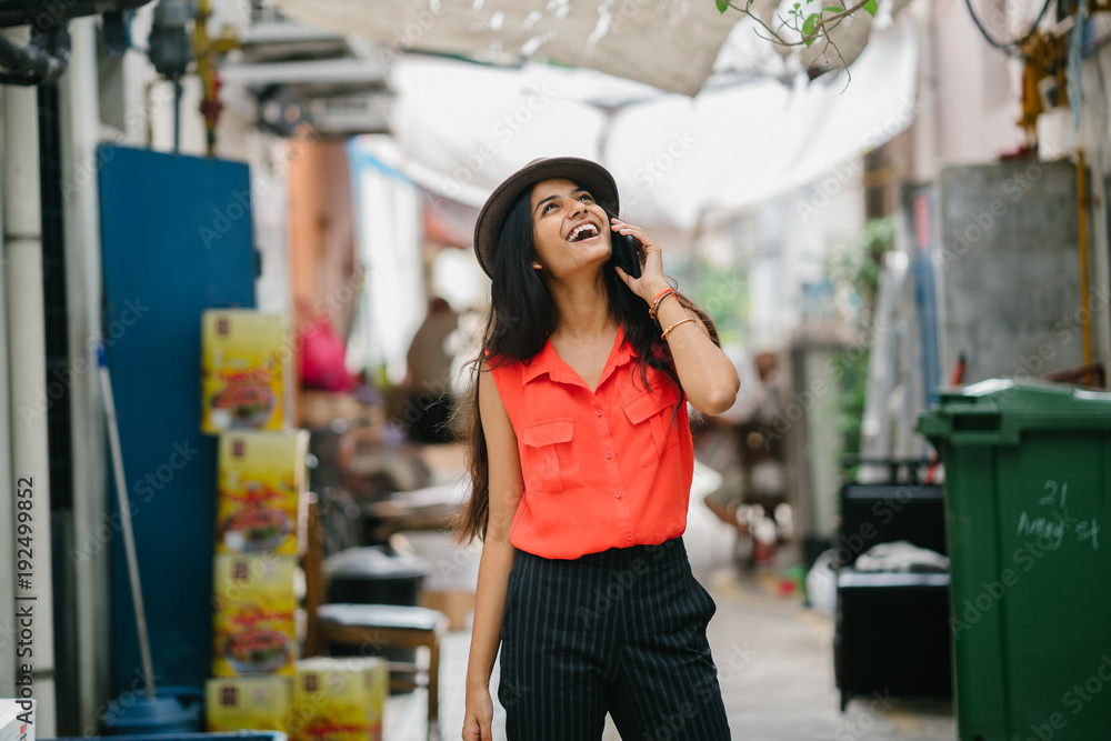 Portrait of a very fashionable and young Indian woman wandering through an alley while on her smartphone talking to someone. She's wearing a casual outfit and a brown fedora hat for the day.