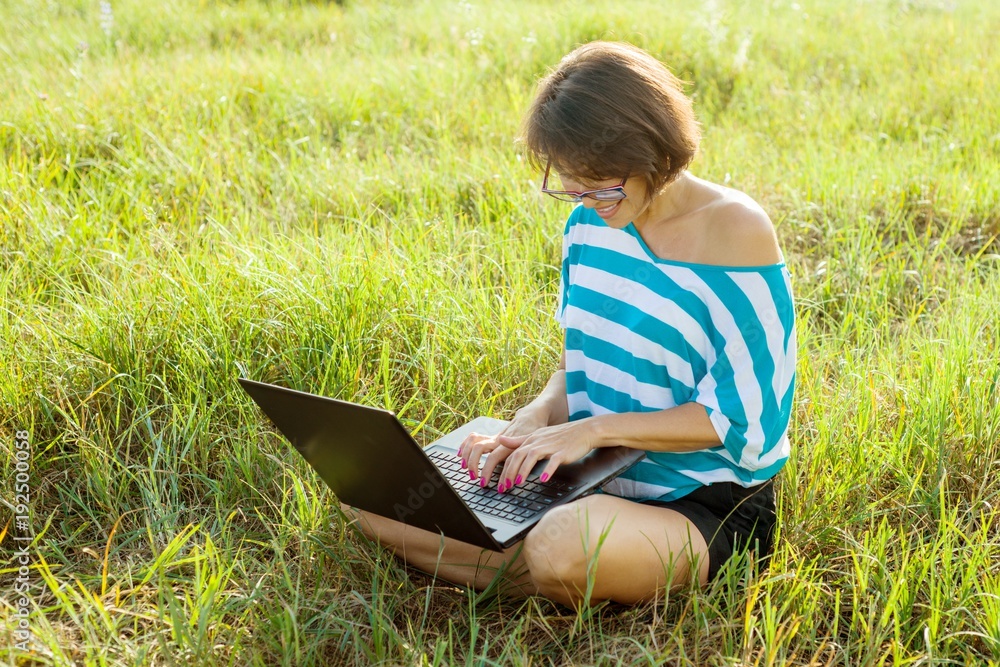 Smiling girl sitting on the grass in bright summer day and working on the computer. Using laptop outdoors, chatting with friends. Smiling girl sitting on the grass in bright summer day and working on the computer. Using laptop outdoors, chatting with friends.