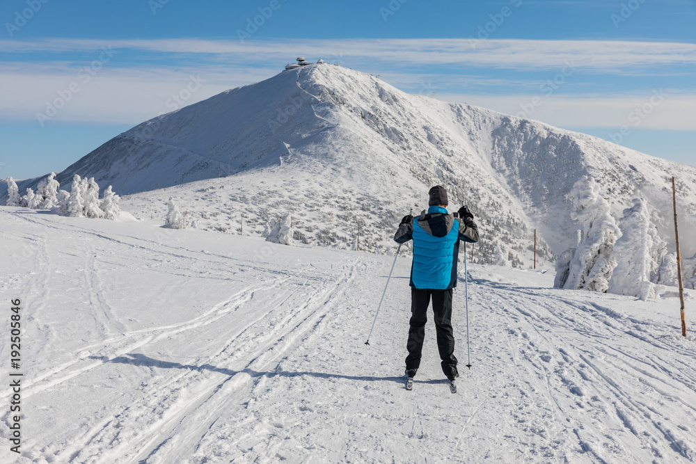 The man in a blue jacket goes on the cross country skiing in the Giant Mountains. In the background is the highest mountain in the Czech Republic Snezka.