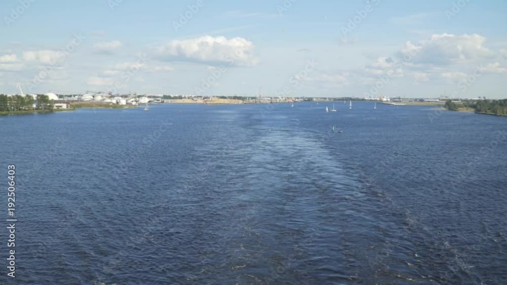 Latvia, Riga Ferry Road Way From the Waves on the Sea Surface After the Ship in River Daugava, City Panorama in the Background. Summer Time Shot Latvia, Riga Ferry Road Way From the Waves on the Sea Surface After the Ship in River Daugava, City Panorama in the Background. Summer Time Shot