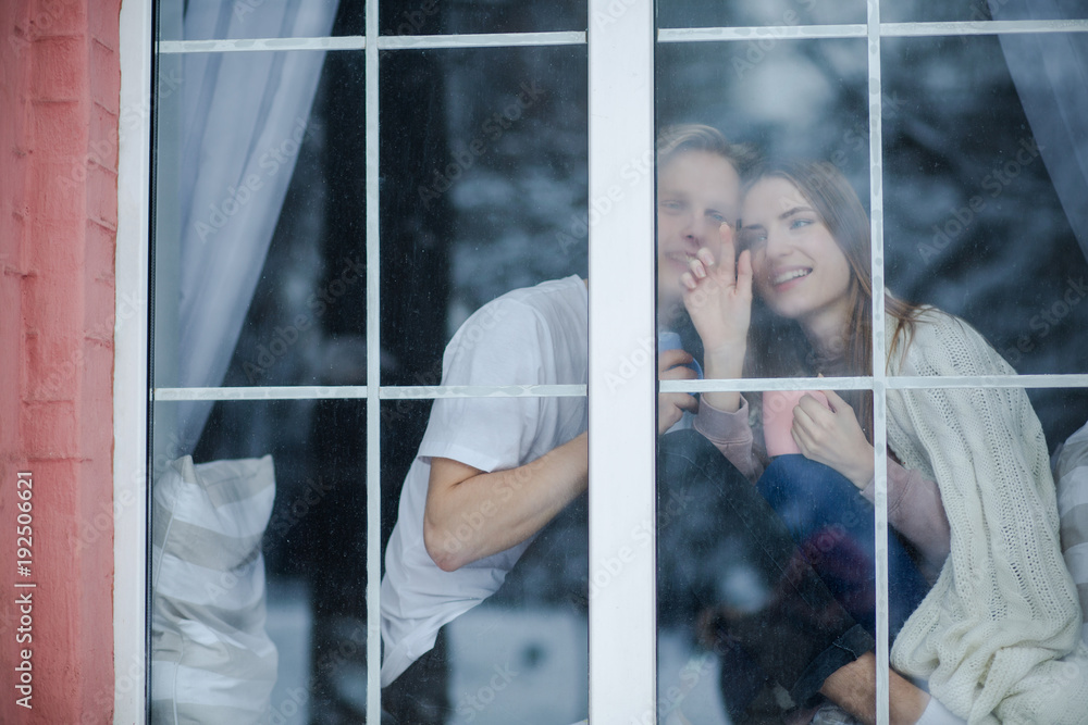 Attractive couple stay at the window. A girl is trying to show a boy falling snowflakes.