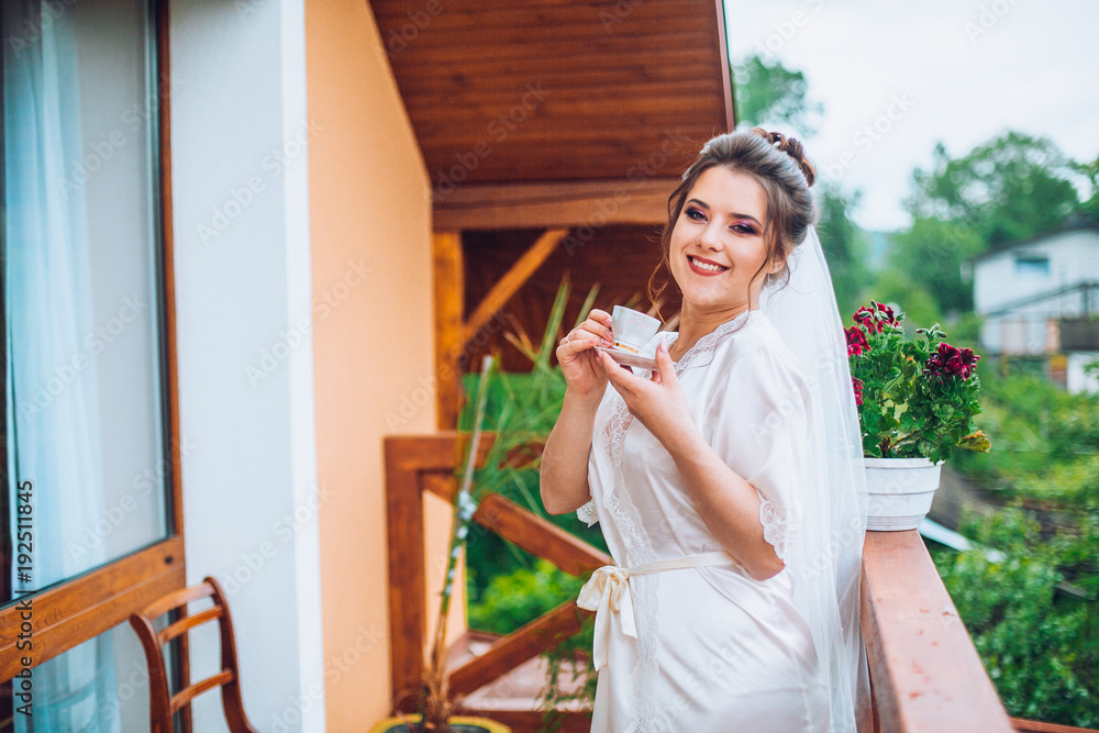 Gorgeous bride in elegant robe drinking coffee in luxury hotel balcony near window, morning before the wedding preparation Gorgeous bride in elegant robe drinking coffee in luxury hotel balcony near window, morning before the wedding preparation