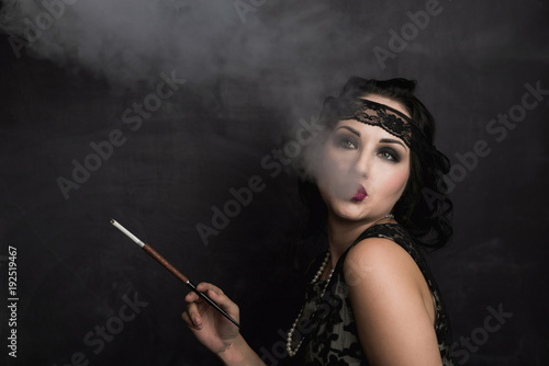 brunette girl in black dress in the style of decadence holds the mouthpiece and blows smoke