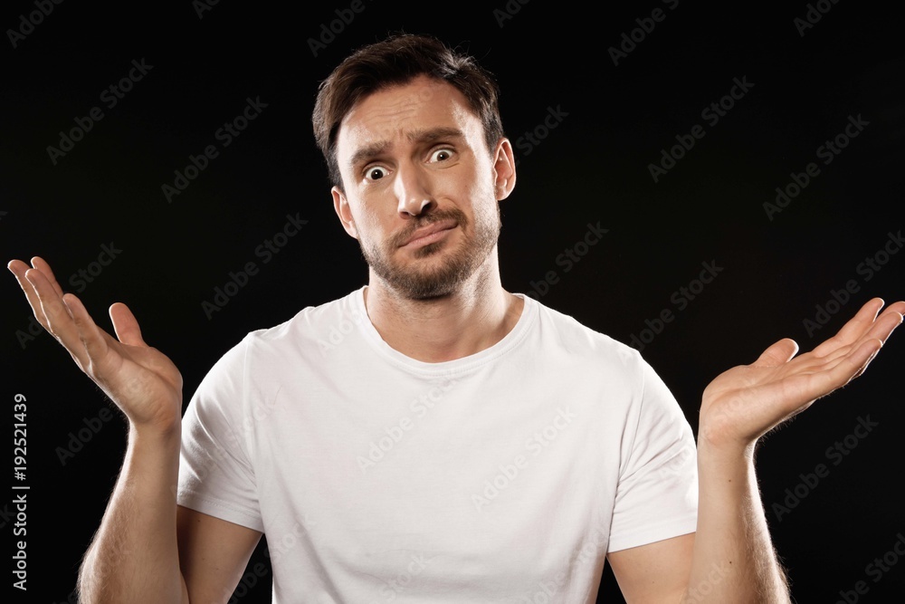 surprised or disappointed young man in a white t shirt cant choose something isolated on a black background. Funny guy is scared with his hands up in wonder surprised or disappointed young man in a white t shirt cant choose something isolated on a black background. Funny guy is scared with his hands up in wonder