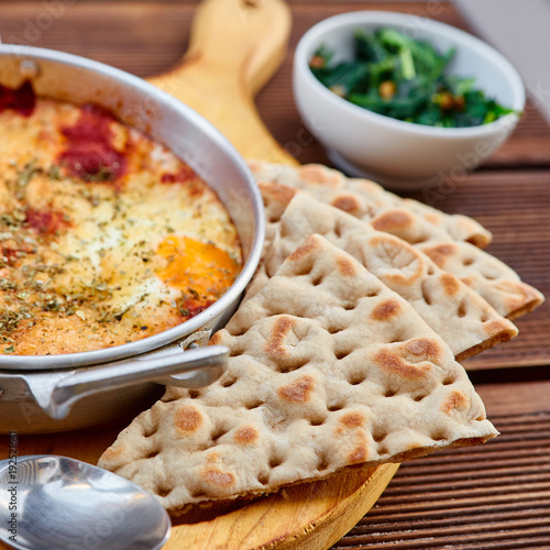 Scrambled eggs with tomatoes, cheese and spices in an aluminum frying pan with handles with homemade thin lavash and juicy greens on a wooden brown table. The photo was taken under natural light. photo