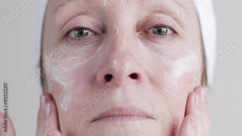 Slow motion shot of beautiful middle-aged caucasian woman in bathroom look at camera and applying cosmetic cream on face. photo