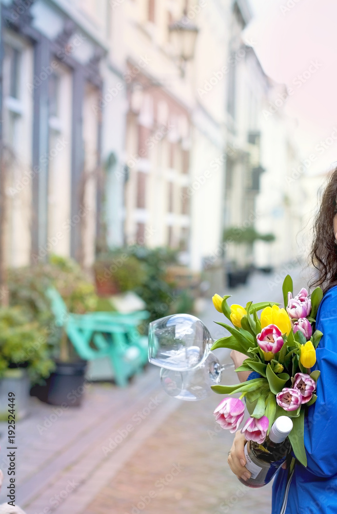 Fototapeta premium girl with a bouquet of spring tulips in pink. In a blue raincoat and yellow boots on the street in Europe. Happy woman. Smile on the face. Card.