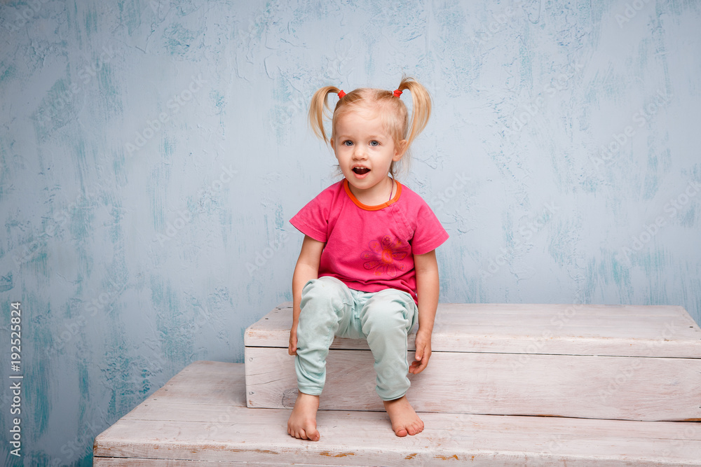 Fototapeta premium Little funny blue-eyed girl child blonde with a haircut two ponytails on her head sitting on a gossip on the background of an old textured wall in blue. Dressed in stylish bright clothes