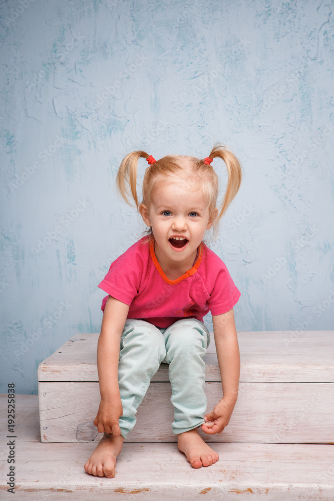 Fototapeta premium Little funny blue-eyed girl child blonde with a haircut two ponytails on her head sitting on a gossip on the background of an old textured wall in blue. Dressed in stylish bright clothes