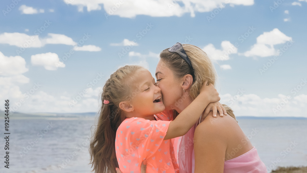 Little cute girl baby and young mother at beach. Mom sits on the shore, and her daughter is coming from behind. The kid hugs and kisses her mother. Mother and her child. Little cute girl baby and young mother at beach. Mom sits on the shore, and her daughter is coming from behind. The kid hugs and kisses her mother. Mother and her child.