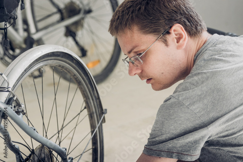 Portrait of a young blond man with short haircut in gray t-shirt with glasses sitting near wheel of broken bicycle