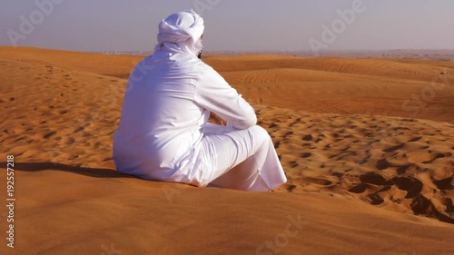 lonely arab man in white kandura sitting in hot sandy desert photo