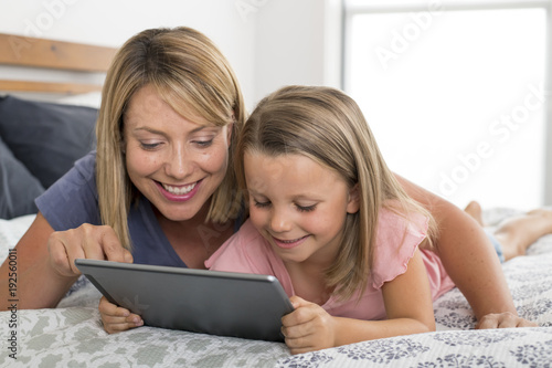  blond Caucasian mother lying on bed with her young sweet 7 years old daughter using internet on digital internet tablet pad together at home