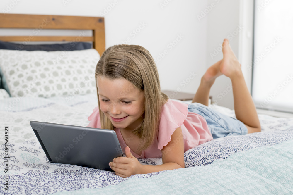 sweet and beautiful blond 6 or 7 years old young girl lying on bed smiling happy using the internet on digital tablet pad watching and having fun sweet and beautiful blond 6 or 7 years old young girl lying on bed smiling happy using the internet on digital tablet pad watching and having fun