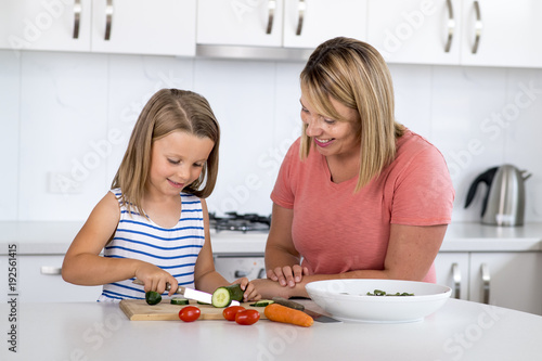 young attractive woman cooking together with her sweet beautiful blond little 6 or 7 years old daughter smiling happy preparing salad