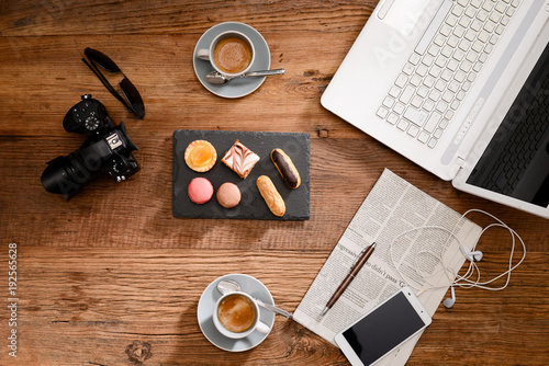 above view still life of two espresso coffee macaroons and french pastries on a rustic wooden tables with a computer, smartphone earphone camera and newspaper