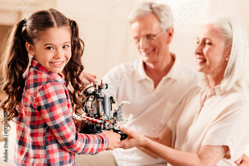 Modern generation. Waist up shot of a preteen kid with curly ponytails looking into the camera with a cheerful smile on her face while visiting her grandparents and showing them a robotic machine.