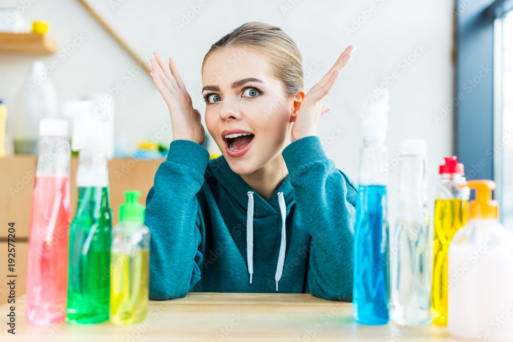 shocked young woman looking at camera while sitting at table with plastic bottles with cleaning products shocked young woman looking at camera while sitting at table with plastic bottles with cleaning products