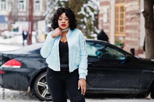 Stylish african american plus size model with mobile phone at hand at winter snowy day against black car.