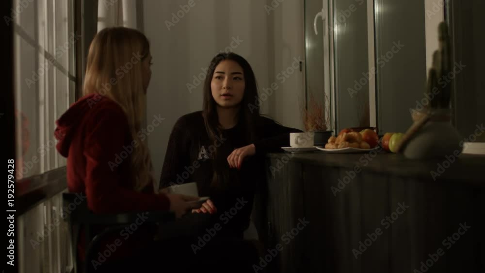 Beautiful multiethnic young women spending evening together, chatting and drinking coffee at home while sitting near window. Positive diverse girls gossiping and enjoying leisure in domestic interior. Beautiful multiethnic young women spending evening together, chatting and drinking coffee at home while sitting near window. Positive diverse girls gossiping and enjoying leisure in domestic interior.