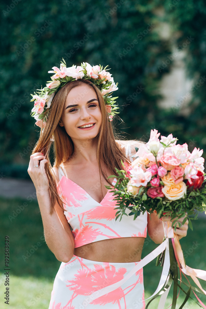 Beautiful young smiling girl in wreath of flowers on her head touching hair with bouquet on green background