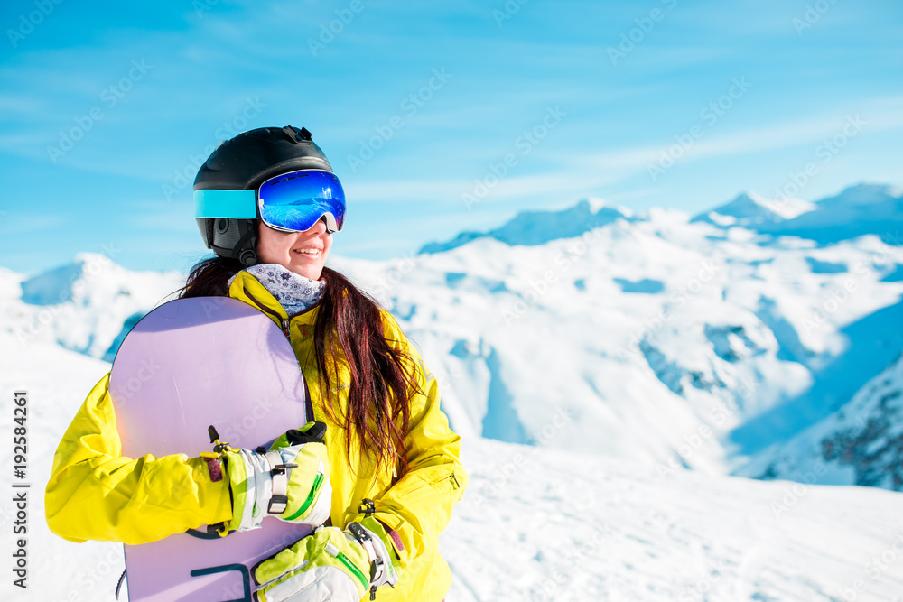 Image of smiling brunette wearing helmet and mask with snowboard on background of snowy hills