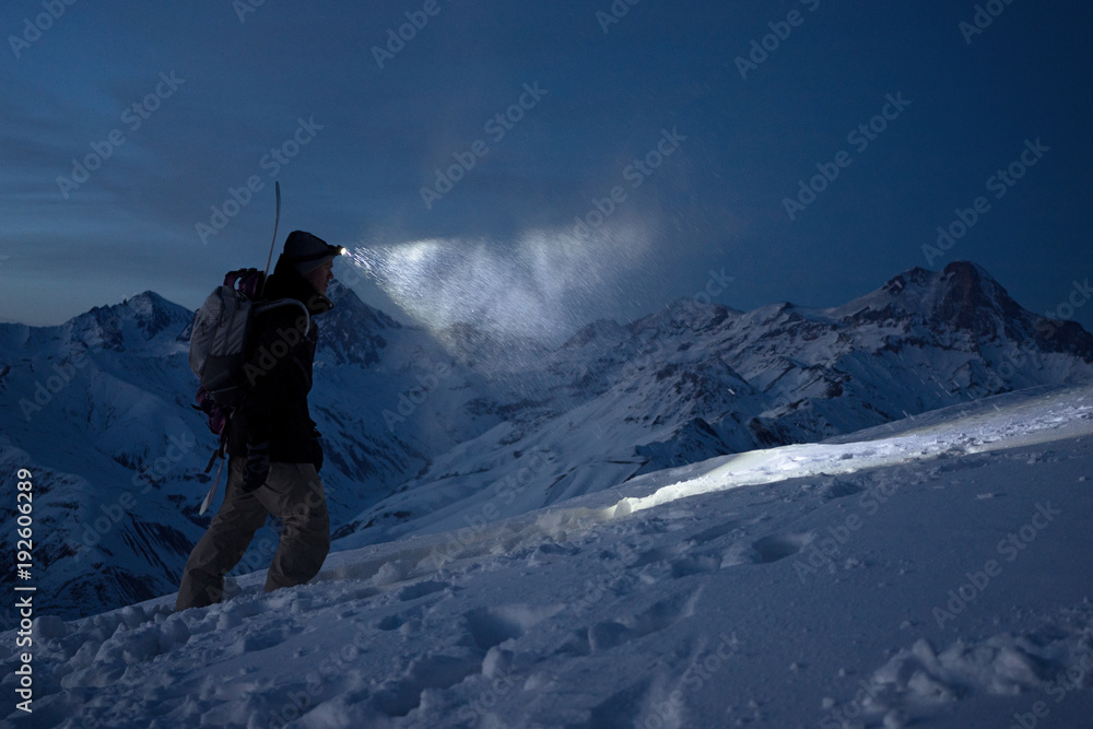 Brave night explorer climbs on high snowy mountains and lights the way with a headlamp. Extreme expedition. Ski tour. Snowboarder commit climb on winter slope. Backcountry - obrazy, fototapety, plakaty 
