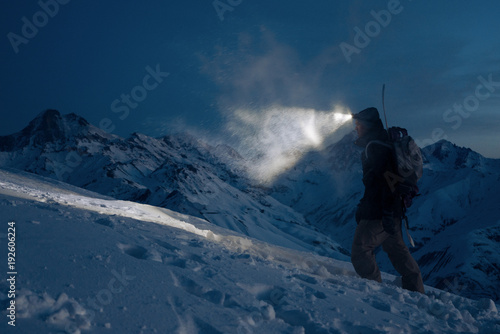 Professional expeditor commit climb on snowy mountains at night and lights the way with a headlamp. Wearing ski wear, backpack and a snowboard behind his back. Backcountry and ski touring