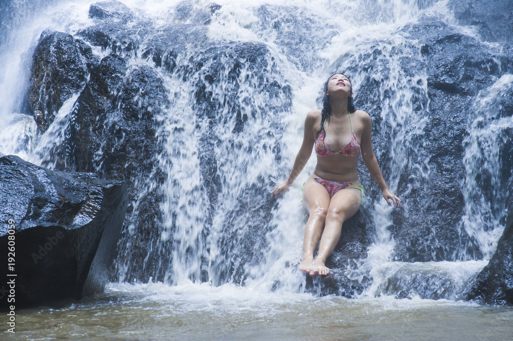 young beautiful and sweet Asian woman in bikini getting body wet under stream of natural amazing waterfall sitting on rock feeling purity young beautiful and sweet Asian woman in bikini getting body wet under stream of natural amazing waterfall sitting on rock feeling purity