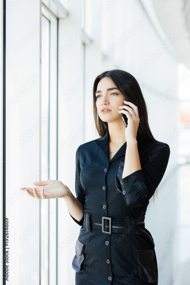 Portrait of young worker speaking using cell phone, looking out the window. Female having business call, busy at her workplace. Portrait of young worker speaking using cell phone, looking out the window. Female having business call, busy at her workplace.