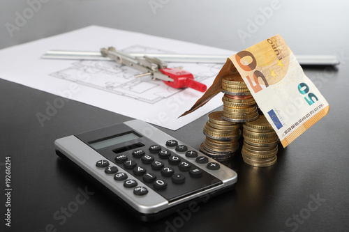 Hausmodell aus Münzen und einer Euro Banknote mit Taschenrechner und Grundrisszeichnung und Zirkel im Hintergrund photo