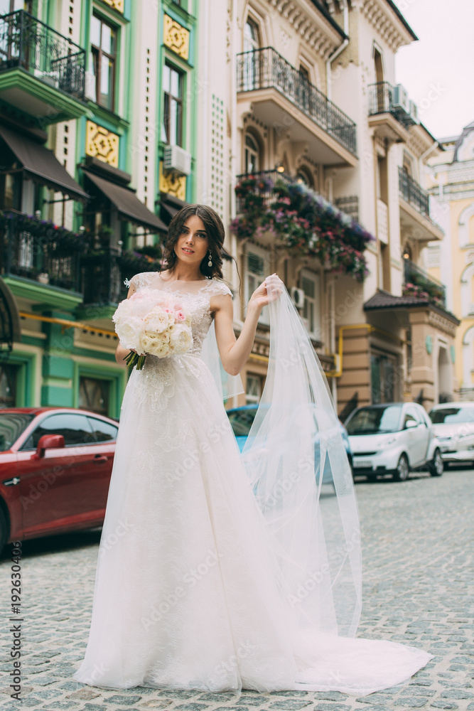 Obraz premium The bride in a wedding dress is standing on the road against the background of a street with a bouquet in her hands and raises her hand
