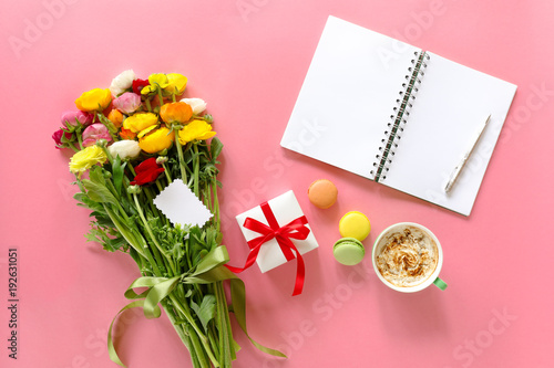 Festive morning concept buttercup flowers bouquet, gift box, cup of cappuccino, makarons cake, clean notebook, pen on the pink background.