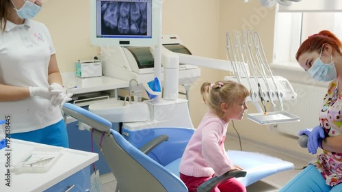little girl came to stomatologist and sits down in dental armchair next to instruments and equipment in bright office