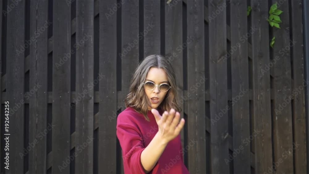 Charming young woman in a red sweatshirt smiling and looking at camera. She is drawing a heart sign and sending an air kiss. A tall wooden fence background. Handheld real time establishing shot Charming young woman in a red sweatshirt smiling and looking at camera. She is drawing a heart sign and sending an air kiss. A tall wooden fence background. Handheld real time establishing shot