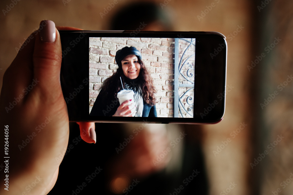 Curly mexican girl in leather cap and plastic cup of coffee at hand on screen of mobile phone.