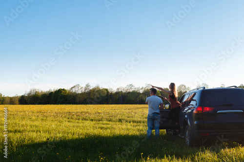 A loving couple watch the sunset, standing leaning against an black car. The young man shows to his girlfriend sunset standing near SUV. Place for text.