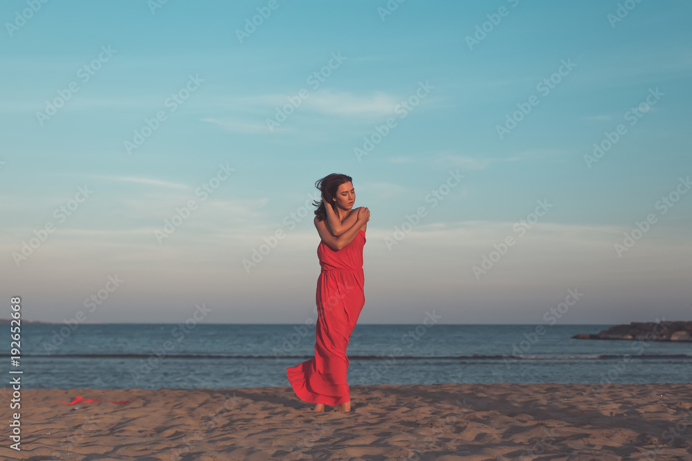 attractive young woman in red dress is standing  on the beach at suset