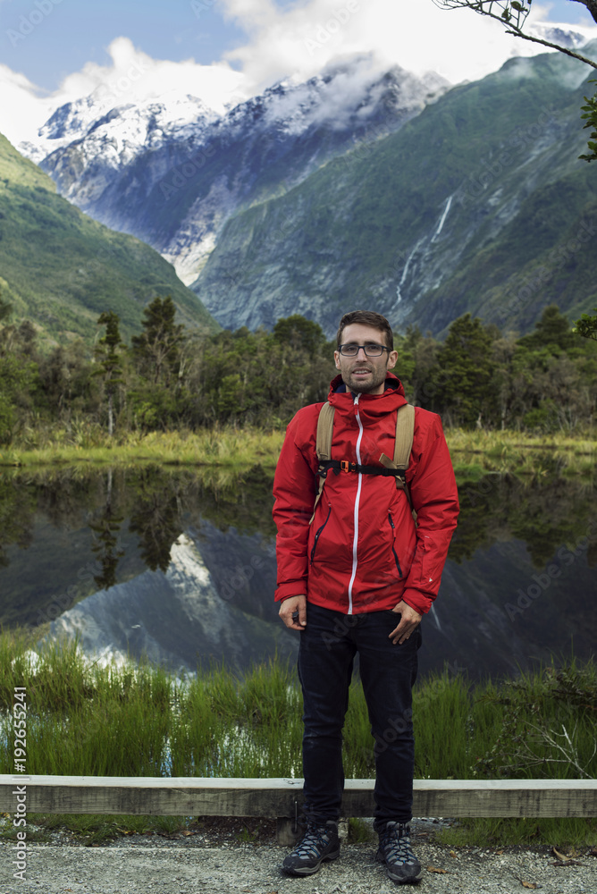 Naklejka premium Hombre joven posando frente a paisaje de picos de montañas nevados y verdes con cielo nublado reflejado en un lago en Nueva Zelanda