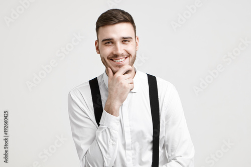 Positive human facial expression, emotions, feelings and life perception. Joyful handsome young bearded guy dressed in stylish white shirt with suspenders, laughing at joke, rubbing his stubble