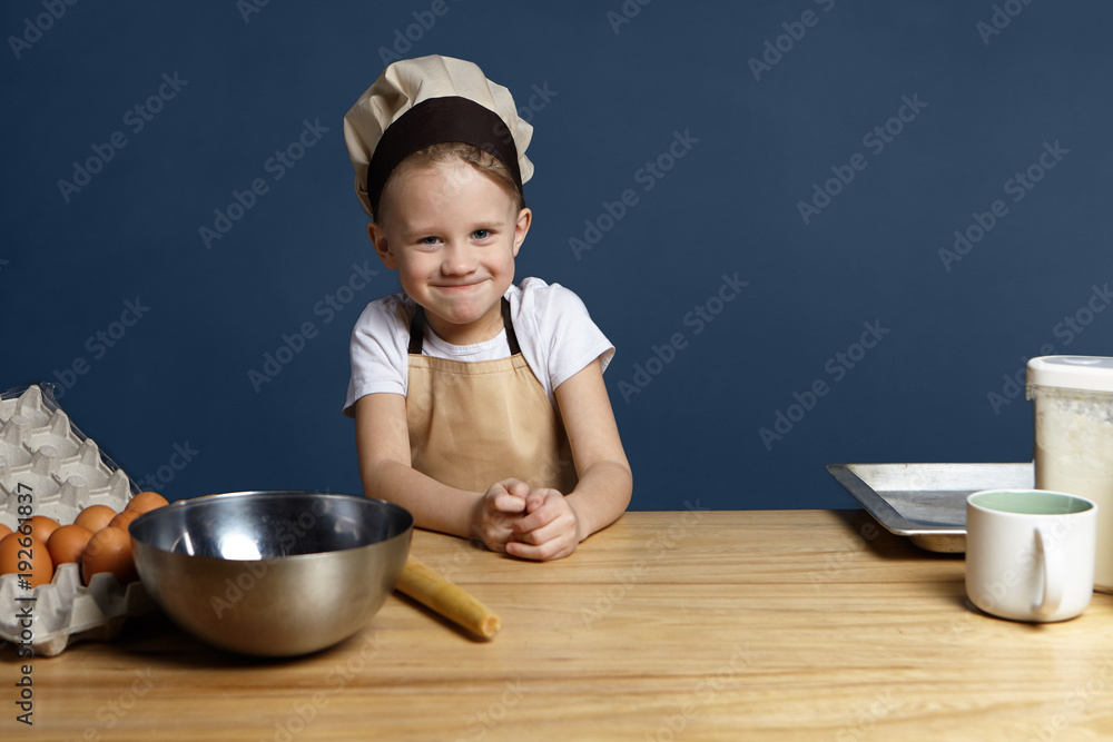 Portrait of funny cute little boy wearing beige apron and chef cap standing in kitchen with metal bowl, cup, tray, eggs and flour on table, ready to make dough for homemade bread, pie or cake Portrait of funny cute little boy wearing beige apron and chef cap standing in kitchen with metal bowl, cup, tray, eggs and flour on table, ready to make dough for homemade bread, pie or cake
