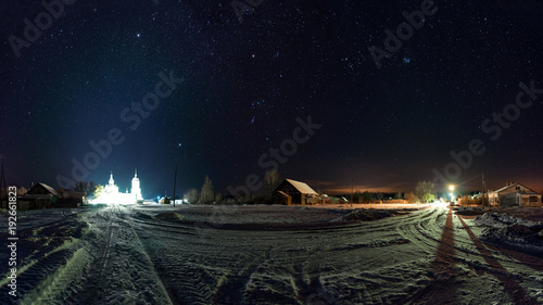 The road in the Russian village. Frosty, winter night. Much snow. Many stars in the sky and the constellation of Orion. The Orthodox Church is illuminated in the distance.
