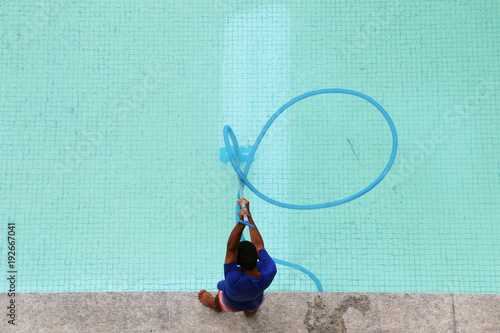 Tablou pe pânză Man is standing on the edge while is cleaning the swimming pool with blue manual