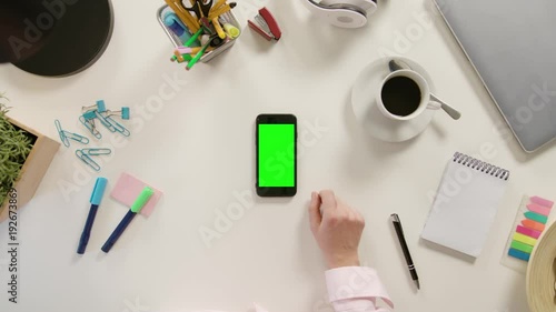A finger touching a smartphone with a green screen. The phone is on the white table. View from the top. Close-up.
