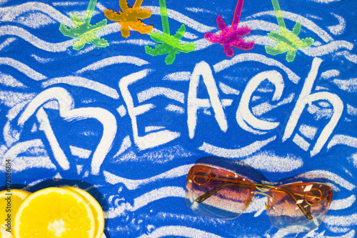 Beach inscription on a blue colored sand with waves, orange and sunglasses, top view, flat lay