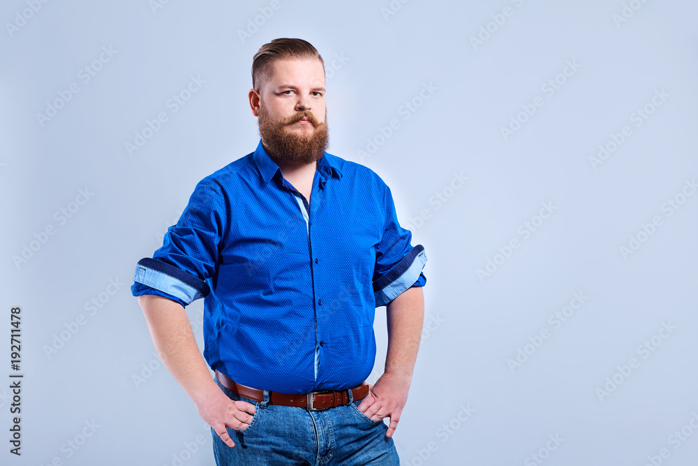 Fototapeta premium A fat, bearded man looks at the gray background with a serious expression of emotion.