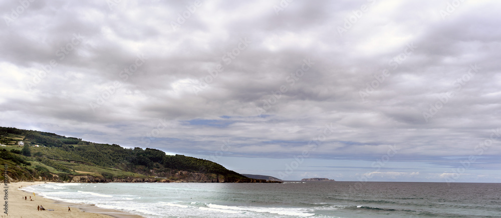 View of the Razo beach on the Atlantic ocean and green mountains to the background on a summer day with many clouds in a landscape typical of Galicia, Spain.