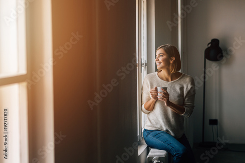 Beautiful young woman drinking coffee and looking through window while sitting at windowsill at home.