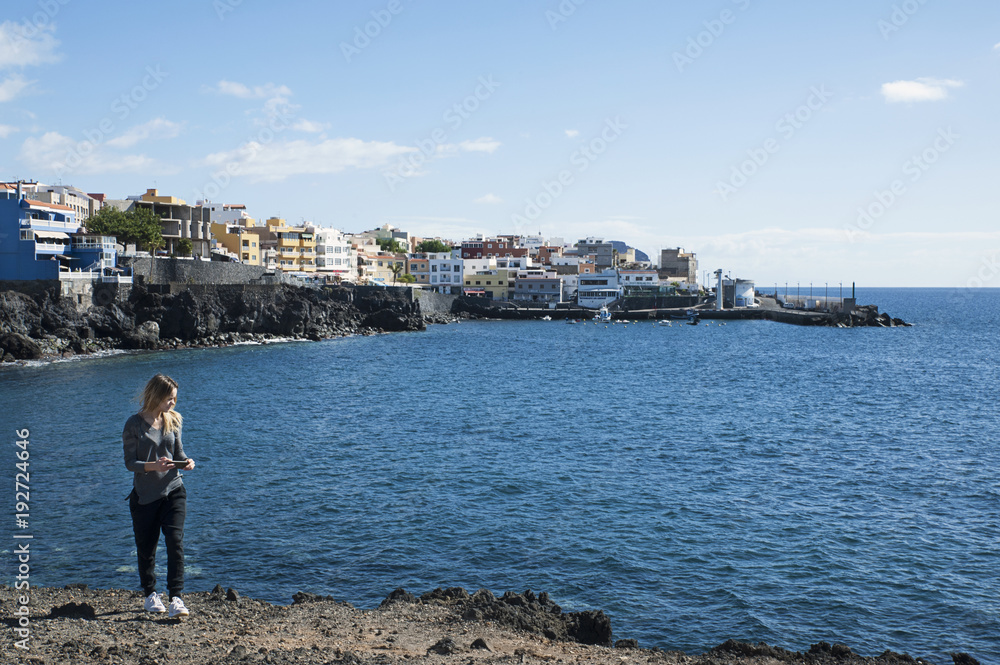 Young millennial female of Caucasian ethnicity holding a phone in the hand and walking away from the coast of Los Abrigos village, in Tenerife, Canary Islands, Spain