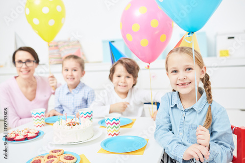 Group of cheerful kids and their teacher in birthday caps holding balloons while sitting by table during celebration